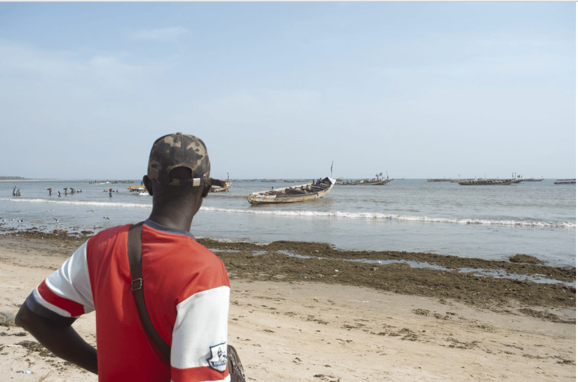 Musa Jammeh’s brother and fellow fisher, Eliman Jammeh, watches other boats return to the beach with fish to sell while his brother’s canoe lies idle (Image: Mustapha Manneh / China Dialogue)